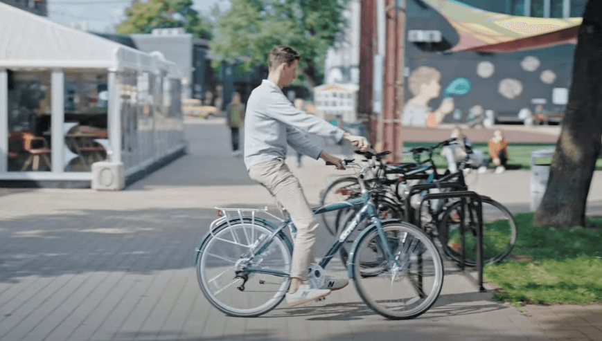 Man riding a blue Oolter Eke electric bike toward a bike rack in a lively urban area, with a mural and people in the background.