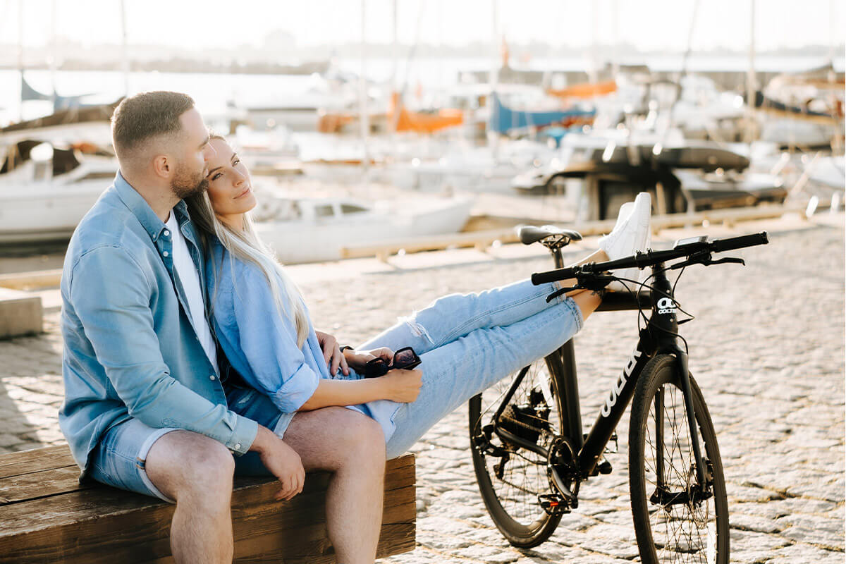 Man and woman sitting on a bench near a harbor, with a black Oolter Torm electric bike beside them, capturing a relaxed moment.