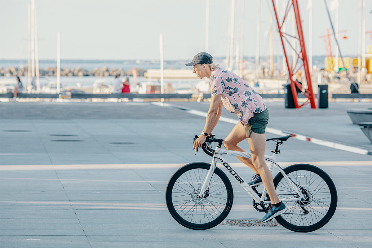 Man in tropical shirt riding a white Oolter Torm S electric bike on a sunny day by the harbor, with boats and cranes in the distance.