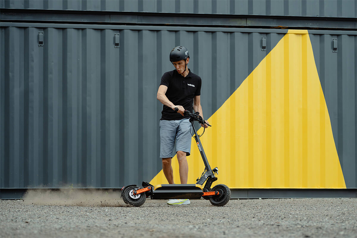 Electric scooter Gpad Storm Max gravel road Image of a man standing still and holding Electric scooter Gpad Storm Max from handlebars while the scooters rear wheel is turning and gravel is flying from the road.