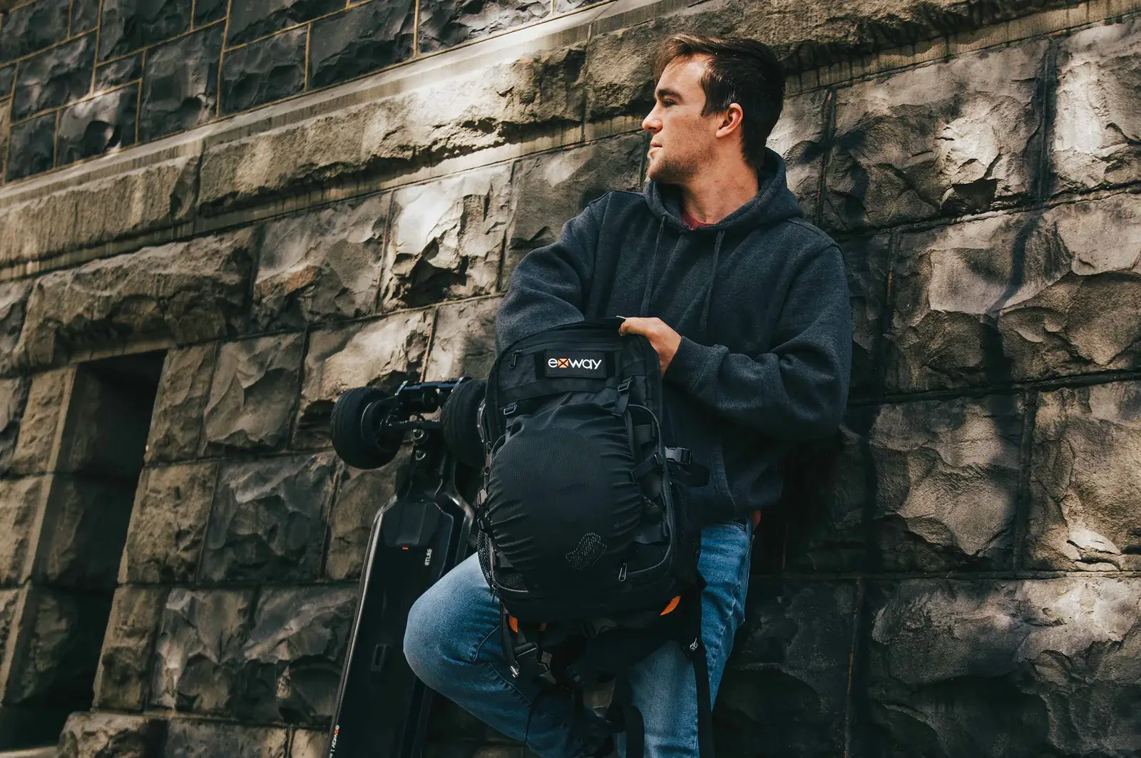 A person leaning against a wall with the Exway Atlas Pro 4WD electric skateboard and an Exway backpack.