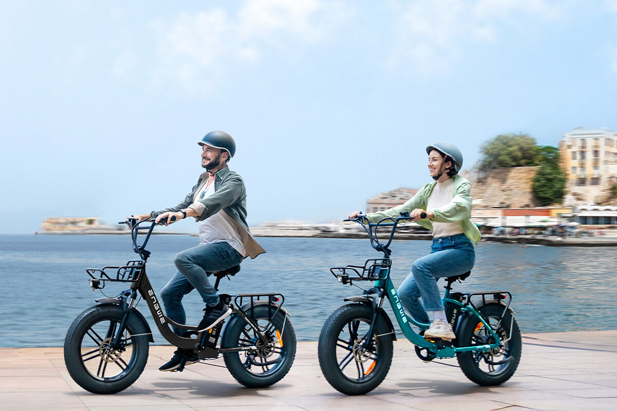 Image of a man and woman riding Electric bicycle Engwe L20 Boost on the sidewalt next to the seaa with a house at the distance.