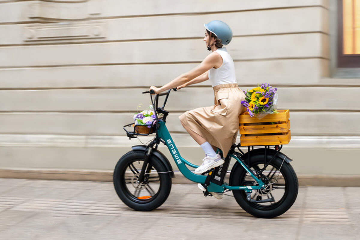 Image of a woman riding Electric bicycle Engwe L20 Boost taht has a bascket at rear carrier and a bag on the fron carrier viewed from left side.