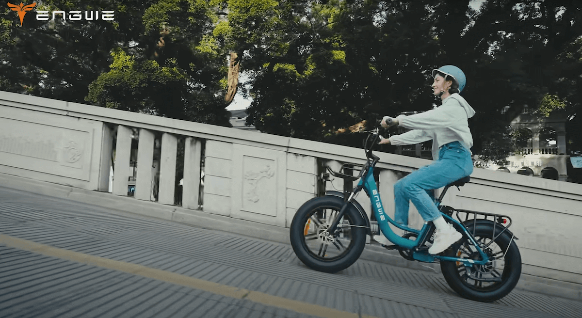 Image of a woman riding Electric bike Engwe L20 Boost uphill on a city road with trees at the background.