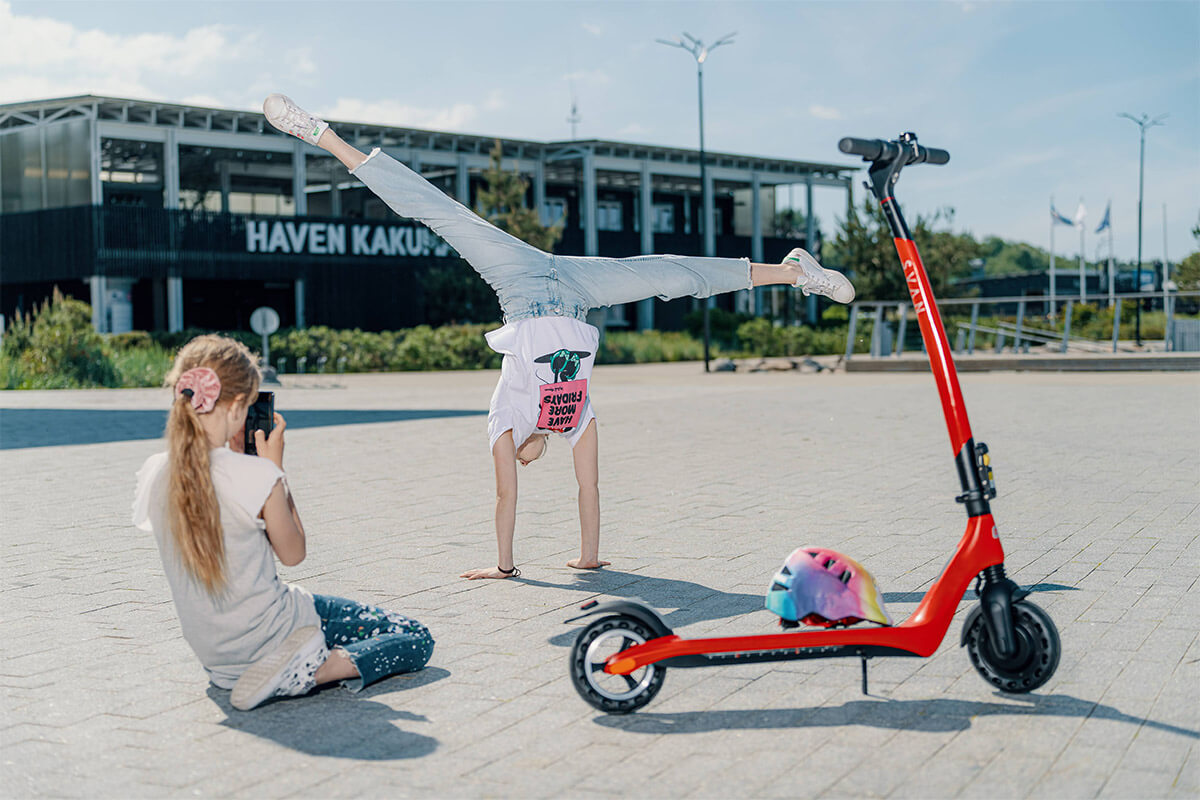 Electric scooter Gpad Svan and girls Image of one teen girl taking. apicture of another girl standing on her hands and red Electric scooter Gpad Svan is standing next to them.