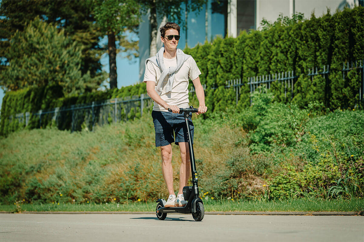 Image of a man riding Electric scooter Gpad Svan Max on summer on the street and plants covered fence is in the background.