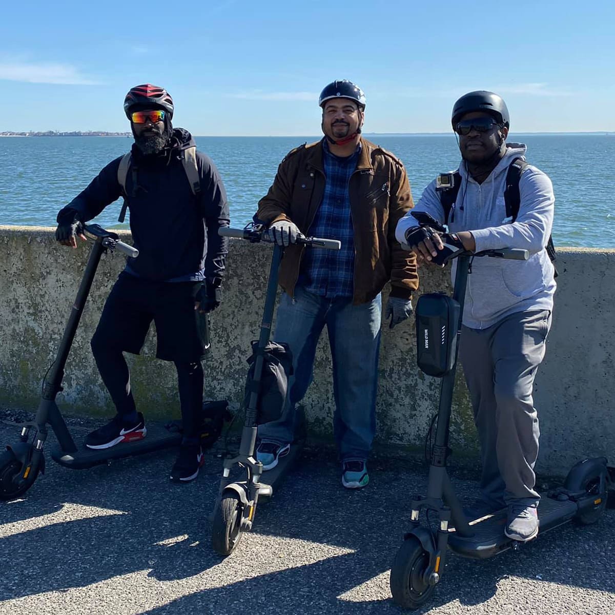 Image of three men standing with electric scooters inmotion s1 on the sidewalk with sea behind them.