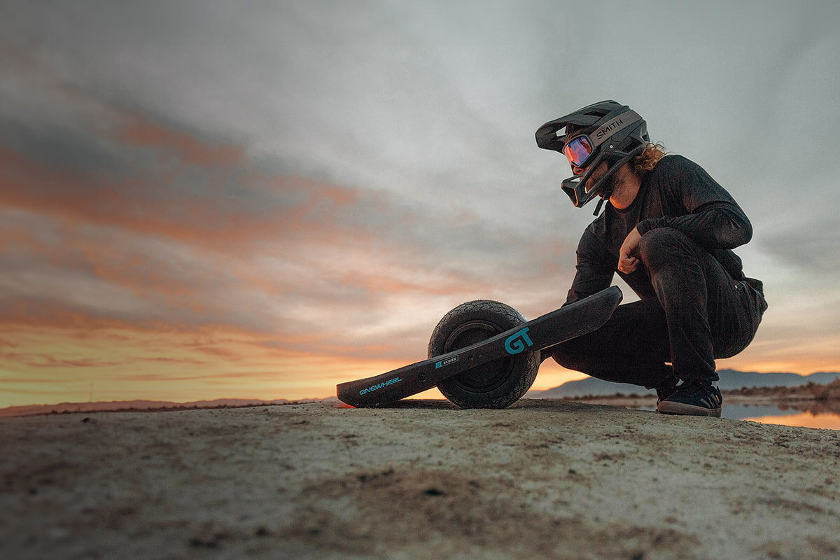 Image of a sunset background and man squatting on the rock and the electric unicycle onewheel gt s rally edition is in front of him.