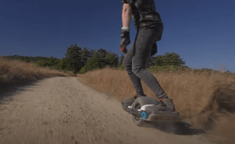 Image of a man riding Electric unicycle Onewheel GT S Rally Edition on gravel road between fields with trees far away.