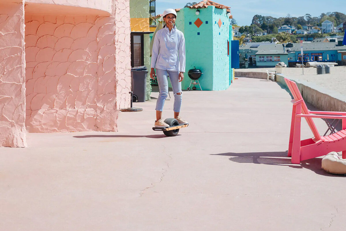 Electric unicycle onewheel pint woman riding Smiling woman riding an Onewheel Pint along a beachside path lined with colorful buildings.