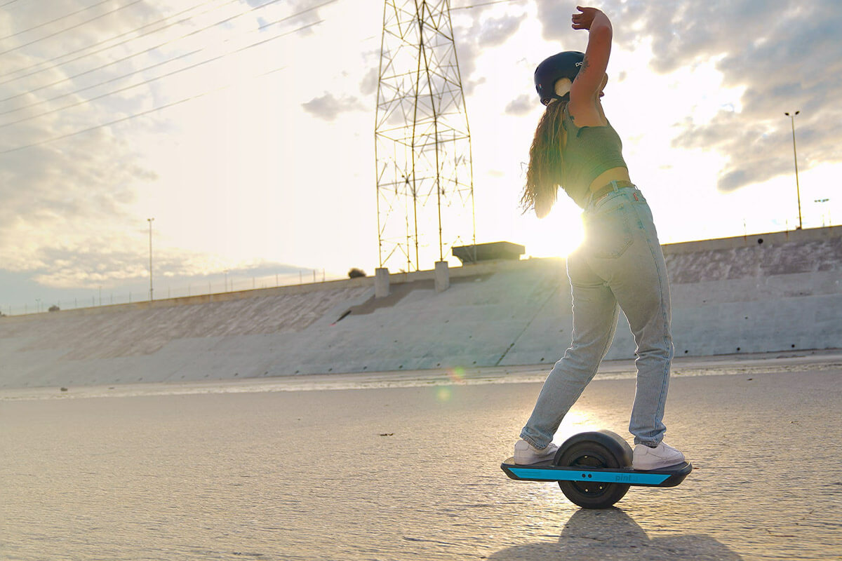 Woman riding an Onewheel Pint X during golden hour with arms raised, captured in front of a setting sun.
