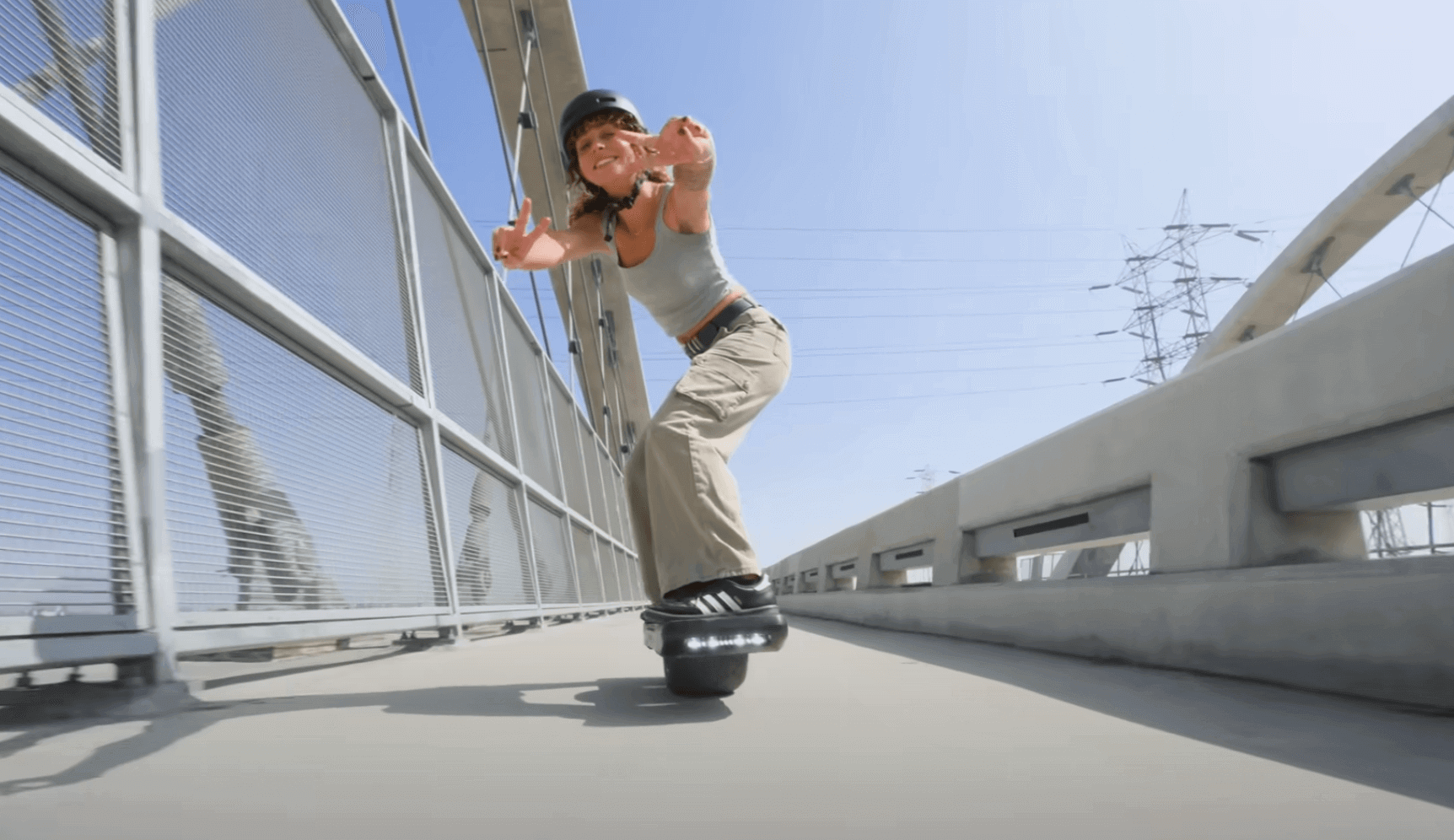 A woman riding Electric unicycle Onewheel XR Classic on concrete bridge towards the camera and smiling and showing thumbs up.