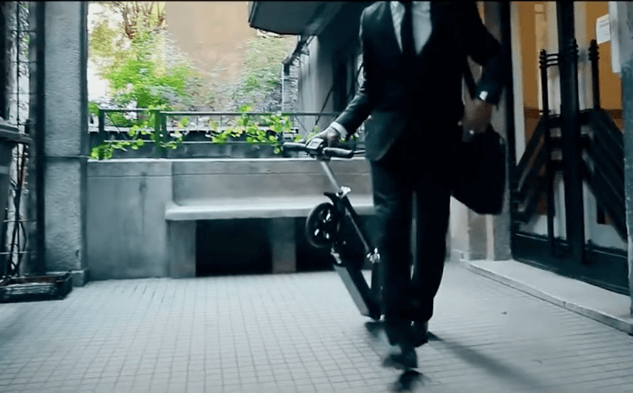 Businessman walking with folded electric scooter E-TWOW S2 Booster V in urban setting, showcasing portability and commuter-friendly design.