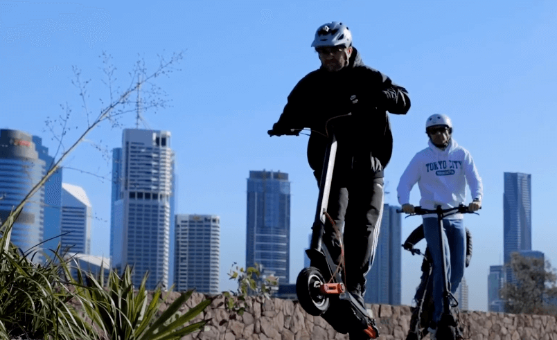 Two riders on Inokim OX Hero electric scooters riding through an urban park with city skyline in background