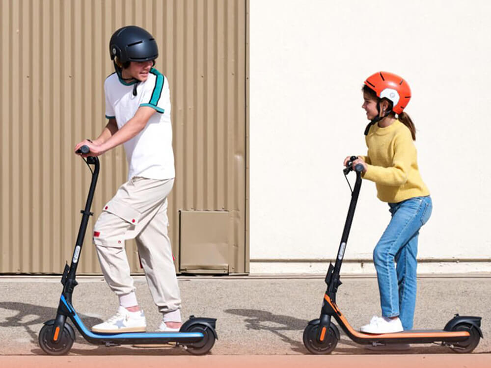 Two kids wearing helmets riding the blue and orange Segway-Ninebot C2 E electric scooters.