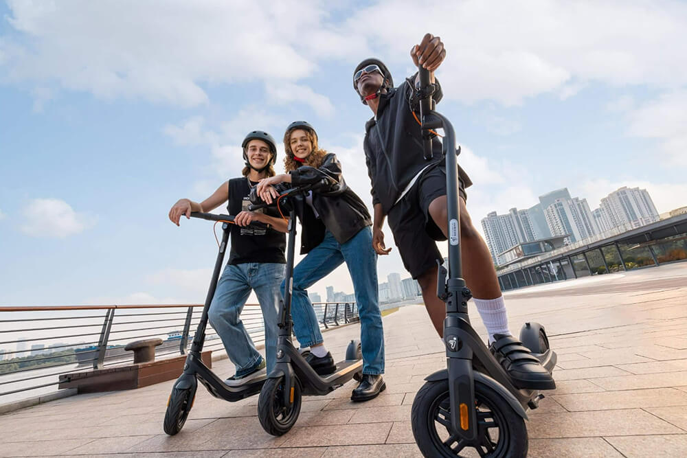 Segway-Ninebot E2 Pro E group ride Trio of riders wearing helmets enjoying a ride on Segway-Ninebot E2 Pro E electric scooters along a riverside promenade, smiling and posing with a modern city skyline in the background under a bright blue sky.