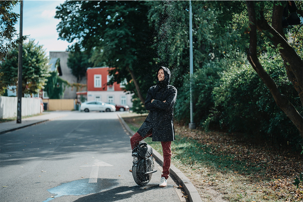 Man in a raincoat standing on a city street with one foot resting on the Inmotion V11 electric unicycle, water droplets falling around them.