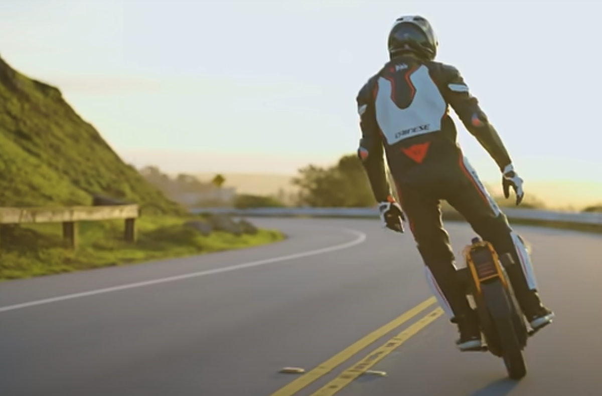 Electric unicycle Inmotion V13 Challenger being ridden on a winding road at sunset by a rider in full protective gear, highlighting high-speed performance and stability.