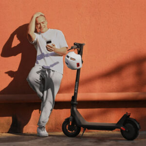 Hymyilevä mies nojaa oranssia seinää vasten, ja hänen vieressään on Xiaomi-sähköpotkulauta sekä kypärä