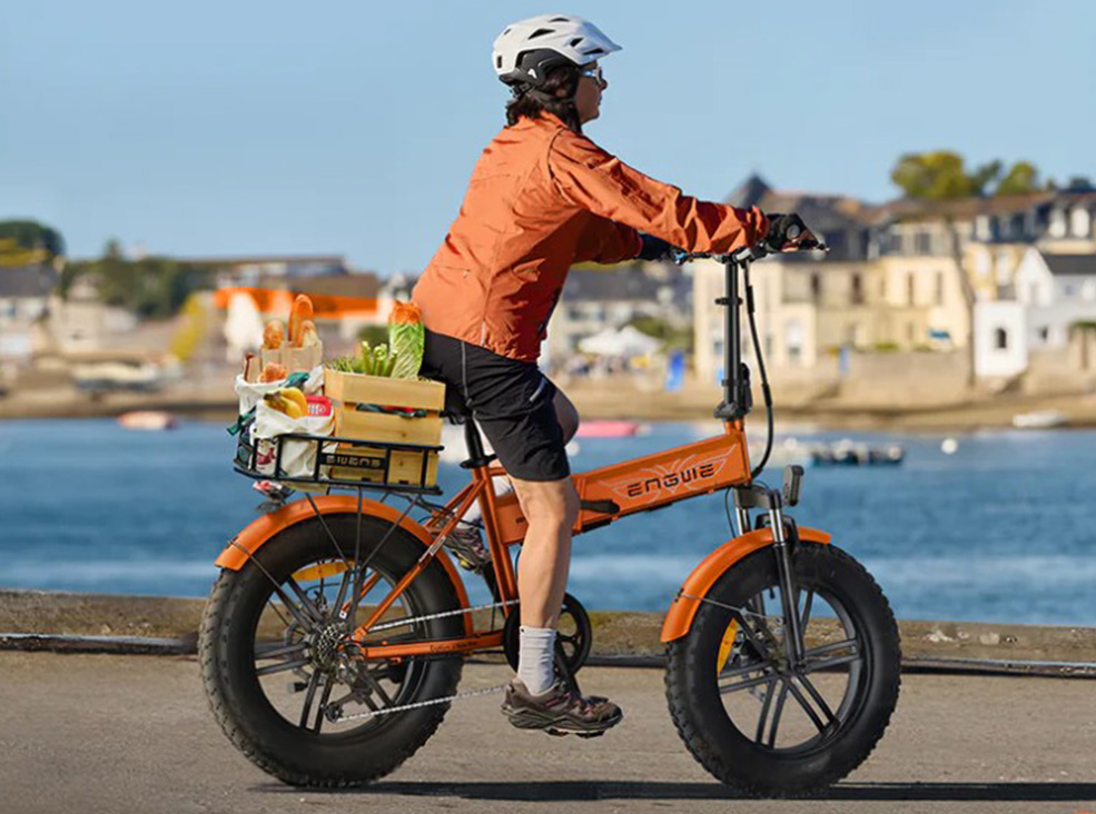 Engwe EP-2 Boost cargo capacity A person wearing a helmet rides an orange Engwe EP-2 Boost fat-tire electric bicycle near the waterfront, carrying a crate of groceries on the rear rack demonstrating the bike's cargo capacity.