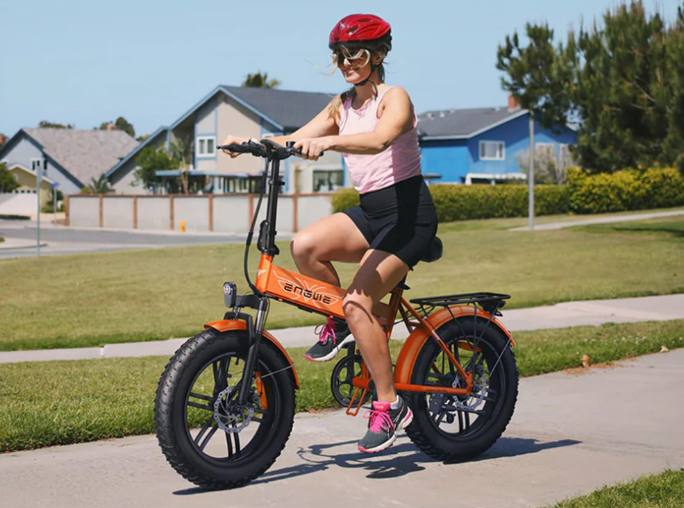Engwe EP-2 Boost girl riding A woman wearing a helmet and sporty clothing rides an orange Engwe EP-2 Boost fat-tire electric bicycle on a sunny suburban path.