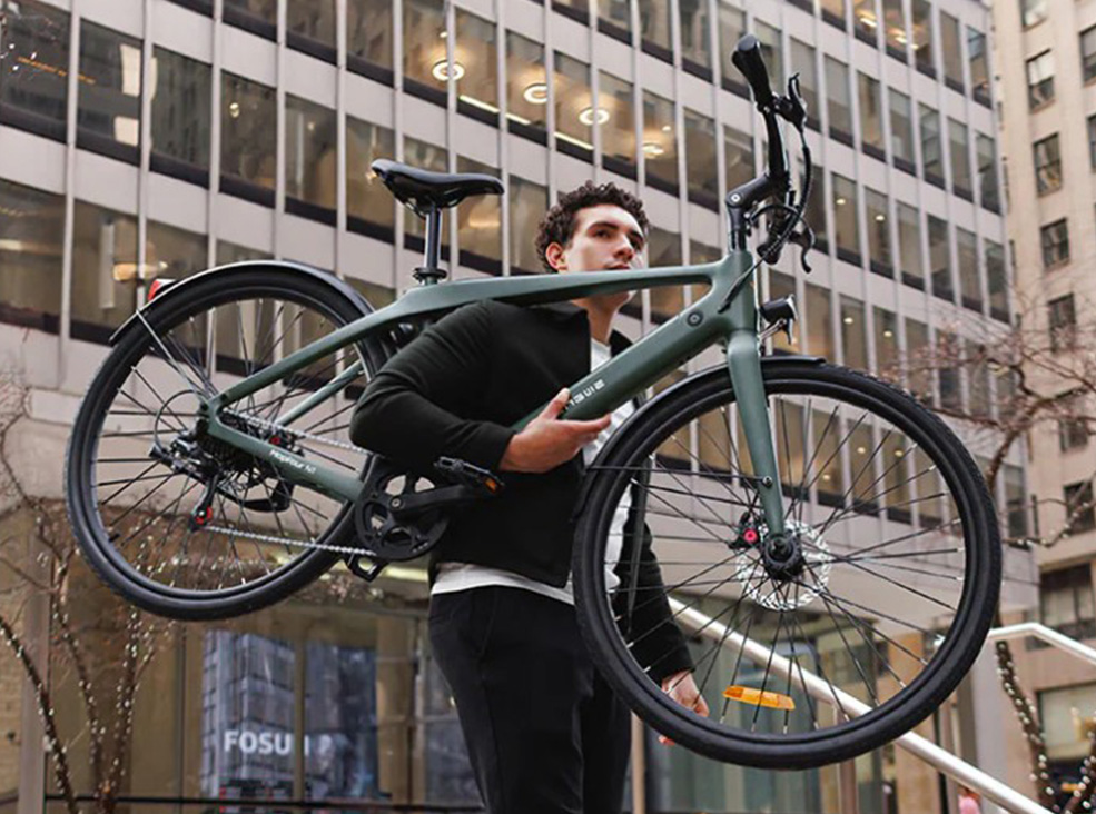 A man carries a lightweight, green Engwe Mapfour N1 Air electric bicycle over his shoulder while walking up city steps surrounded by tall office buildings.