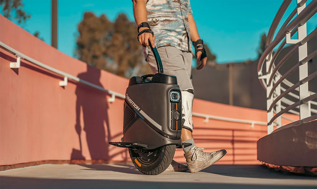 A rider walking along a sunlit ramp while pulling the Inmotion V9 electric unicycle by its extendable trolley handle, with protective gear on their legs and arms.