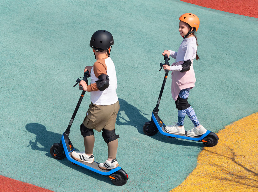 Segway-Ninebot C2 Lite Kids lifestyle Two kids wearing helmets and protective gear riding the Segway-Ninebot C2 Lite Kids electric scooters.