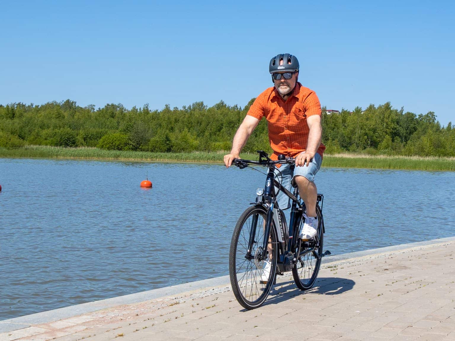 Tunturi eMotion man riding3 A man wearing a helmet and sunglasses rides a black Tunturi eMotion e-bike along a waterfront path on a sunny day.