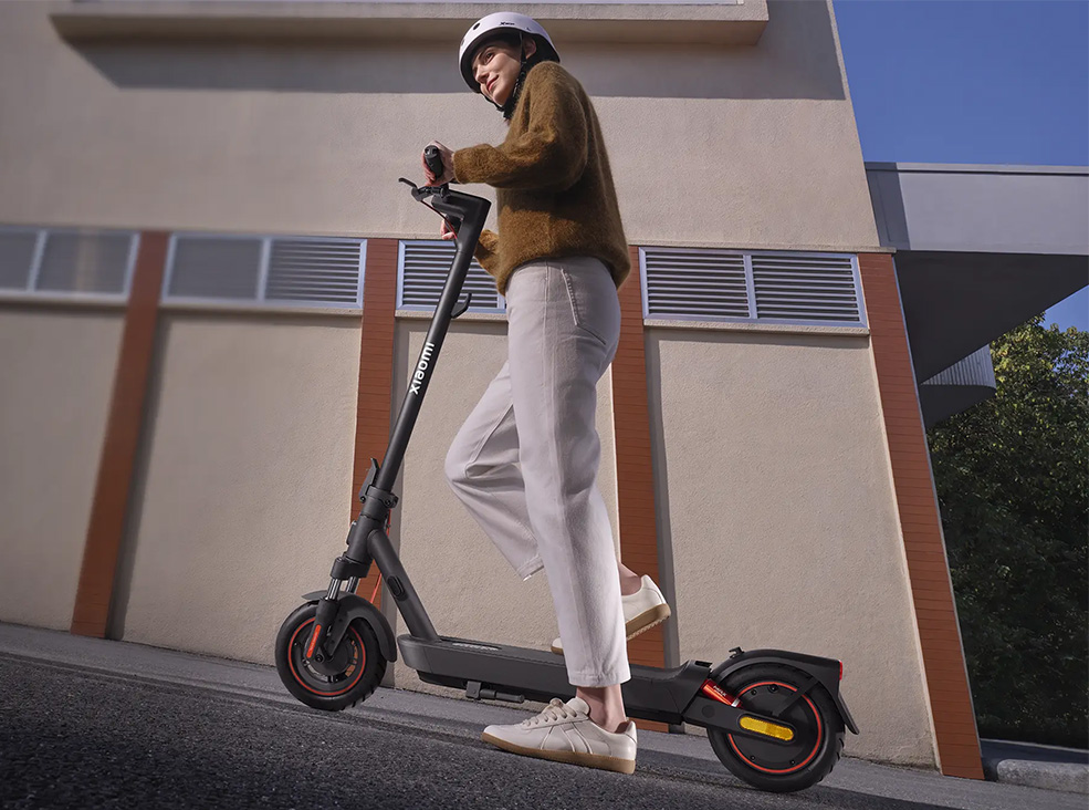 Person wearing a helmet riding the Xiaomi 5 Max electric scooter uphill demonstrating the scooter's slope parking system on a city street in daylight.