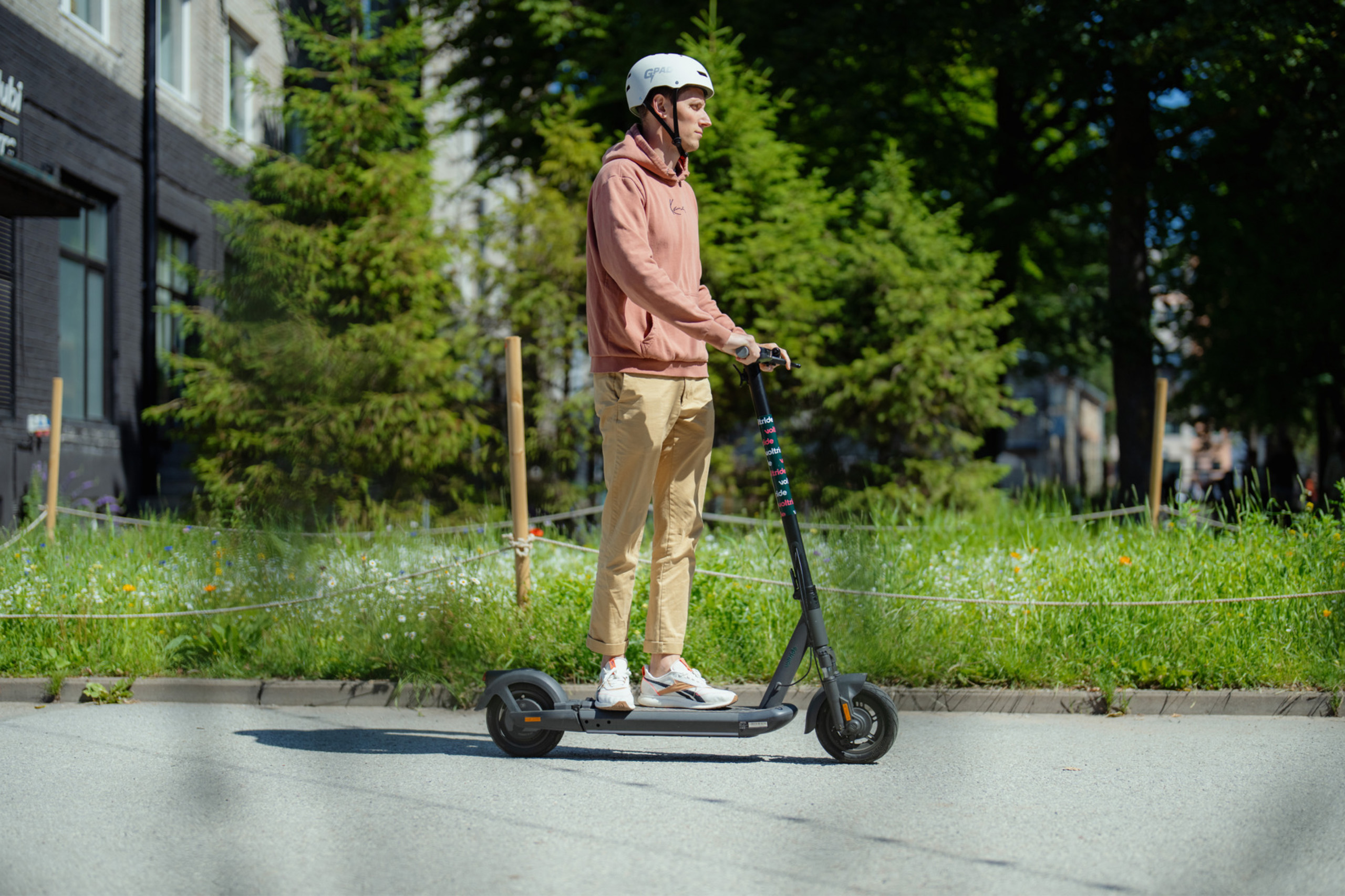 A person wearing a helmet and casual clothes rides an electric scooter along a quiet street bordered by greenery and trees.