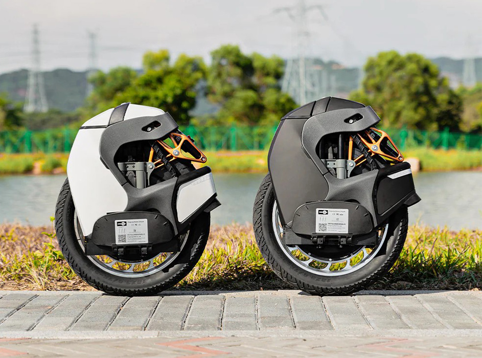 Two Kingsong S18 Pro+ electric unicycles, one white and one black, are parked side by side outdoors, showing their suspension arms, treaded tires, and compact body design with a lake and trees in the background.