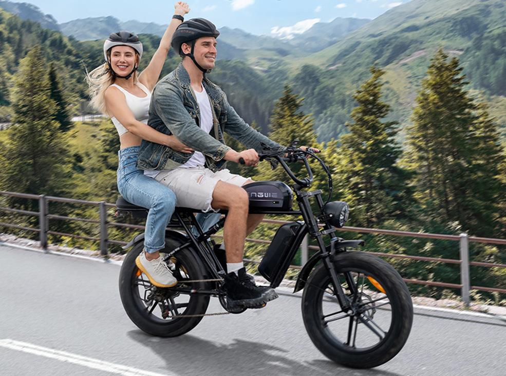 engwe-m1-lifestyle1 Two people riding the ENGWE M1 black fat-tire electric bike on a mountain road, both wearing helmets, with scenic forested hills in the background.