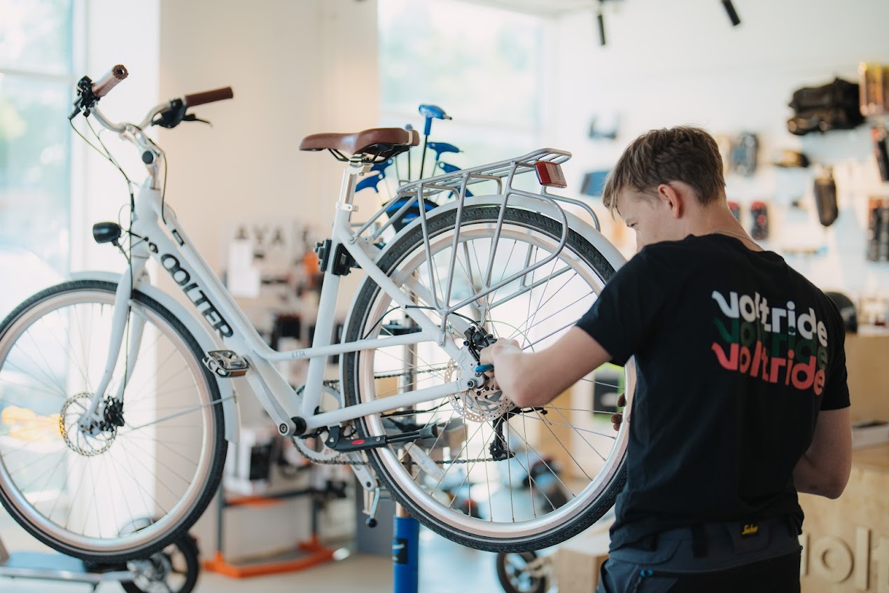 A bicycle mechanic adjusting the rear wheel of a white city bike mounted on a repair stand in a bright workshop