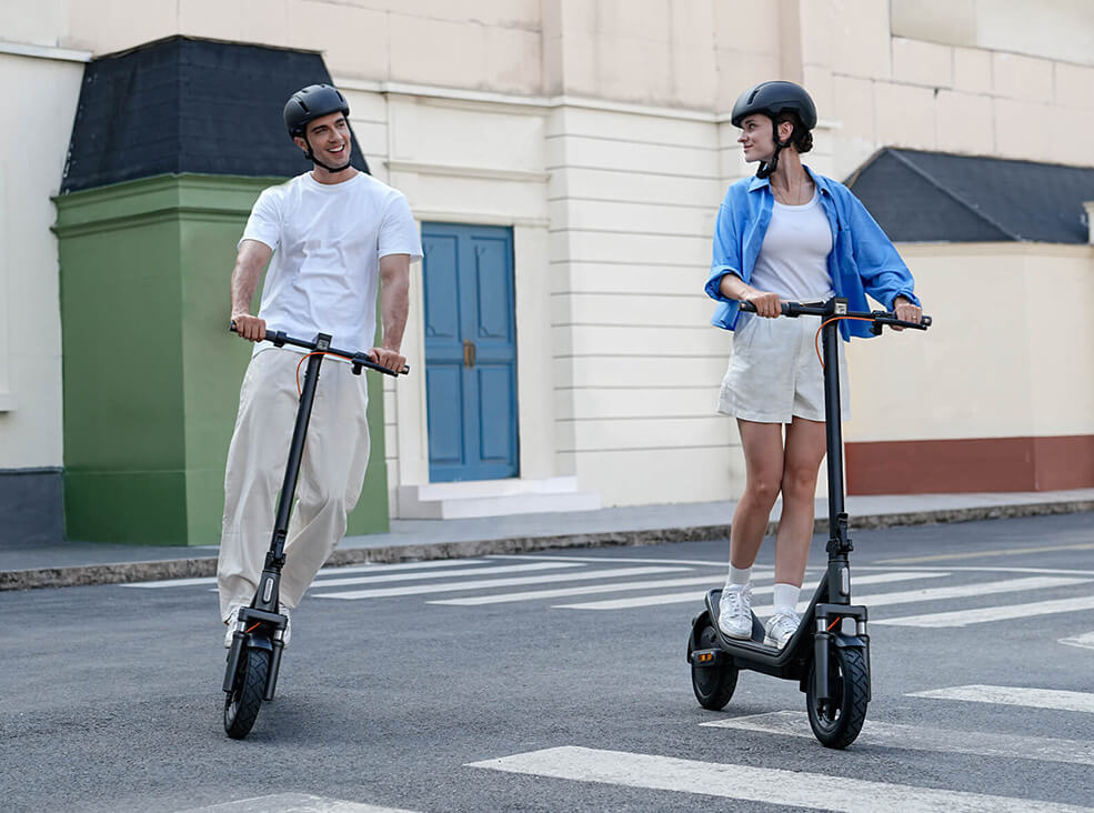 Two people riding Xiaomi 6 electric scooters through a city street while wearing helmets and casual clothing.