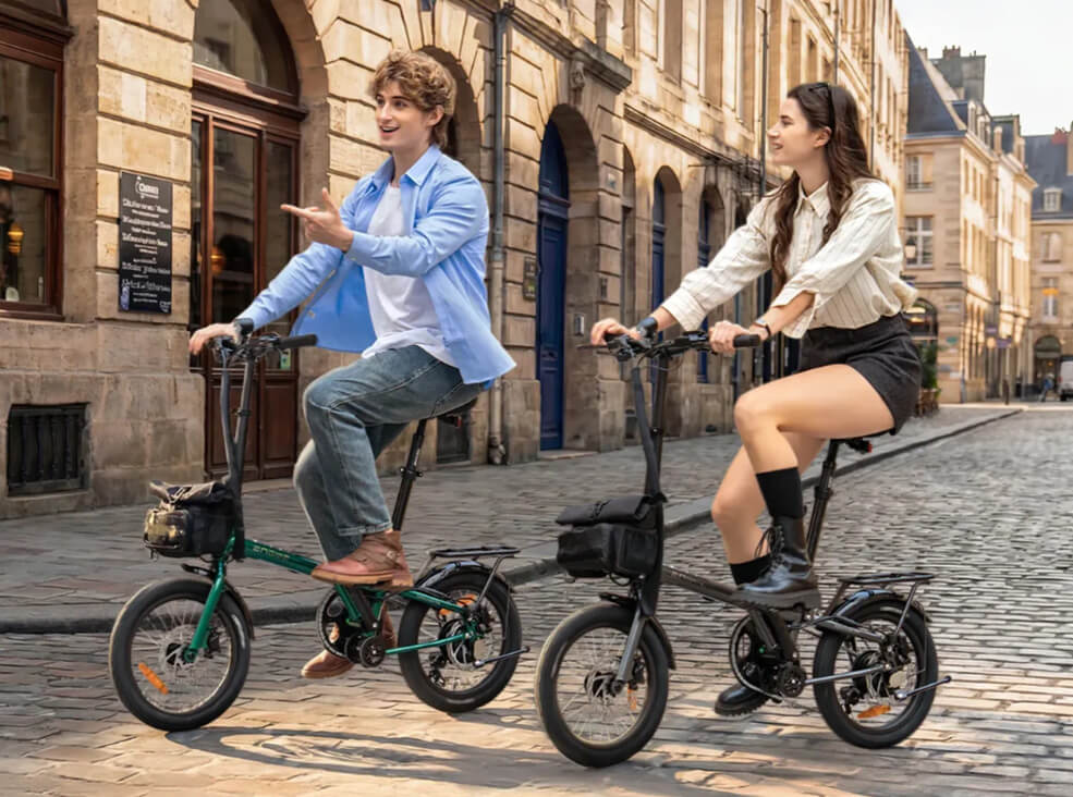 Two people riding the Engwe Zip compact folding electric bicycles on a cobblestone street in a historic European city center, with stone buildings and urban architecture in the background.