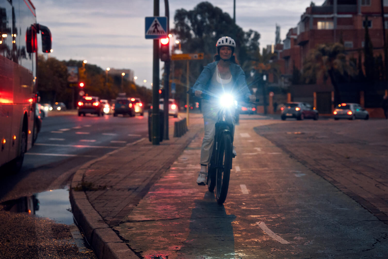 Riese Müller Charger 5 Mixte lifestyle2 Cyclist riding a Riese & Müller Charger5 Mixte electric bike on a city bike lane at dusk, with a bright integrated front light, urban traffic, and wet pavement reflecting evening lights.