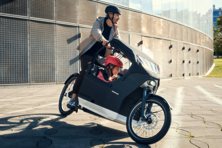 Man riding a Riese & Müller electric cargo bike with a helmeted child seated under the front canopy on a sunny city street.
