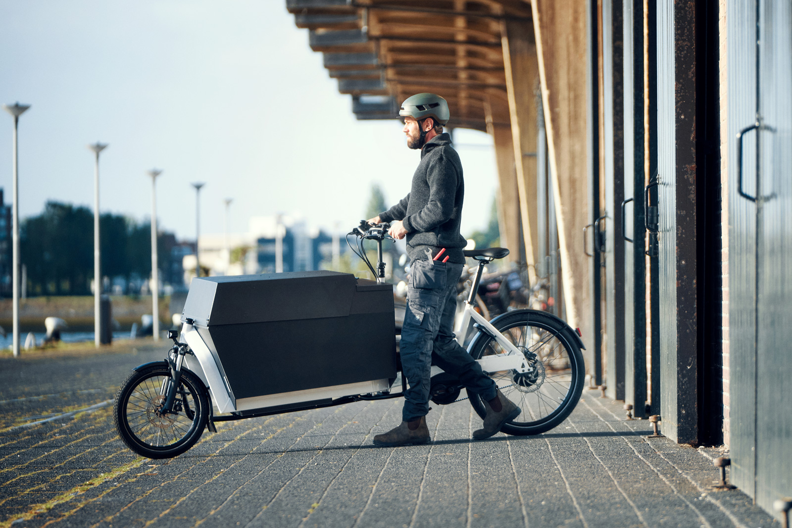 Helmeted man in workwear pushing an electric cargo bike with a large enclosed front cargo box outside an industrial building.
