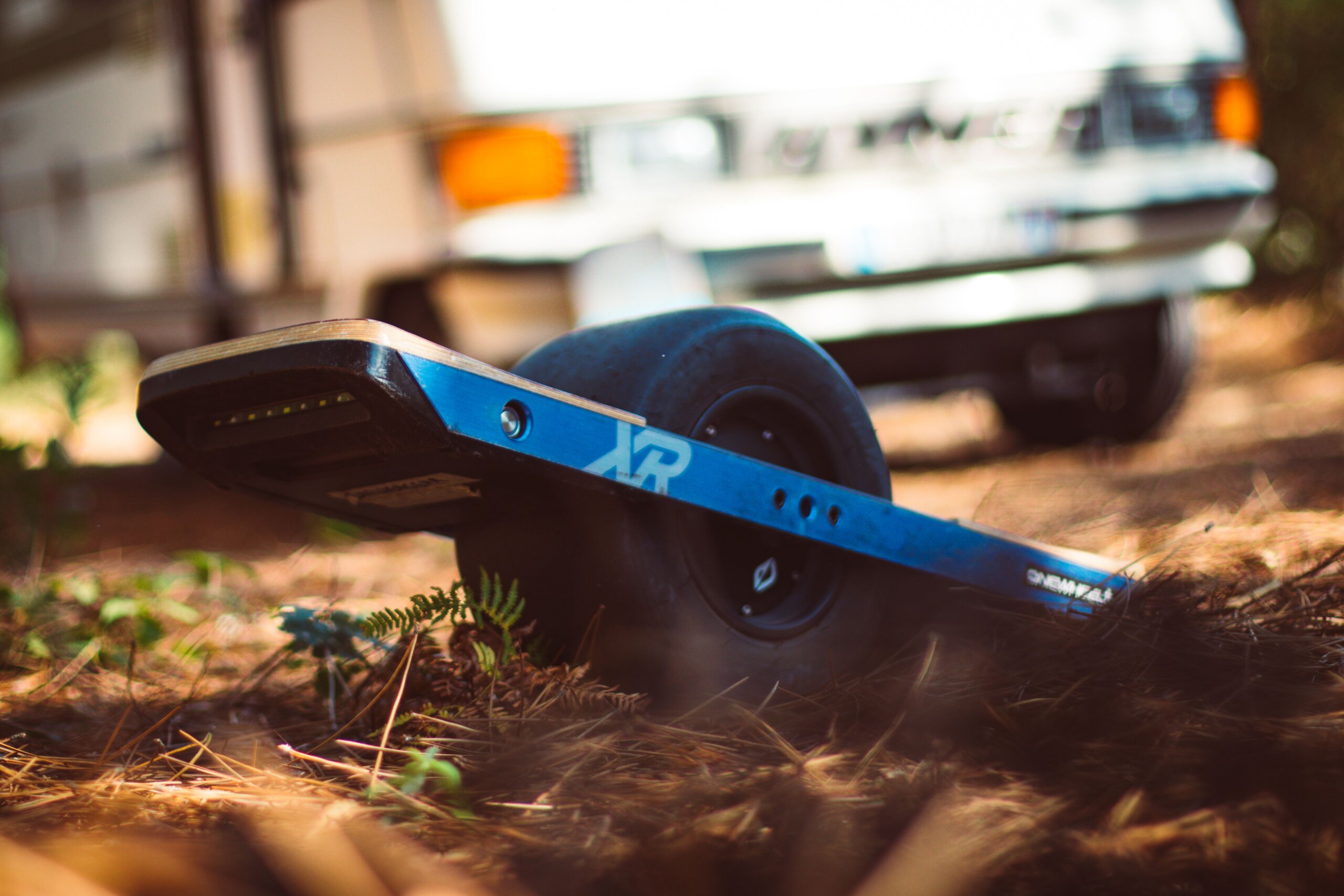 Close-up of a blue Onewheel electric board resting on a forest floor with a camper van blurred in the background.