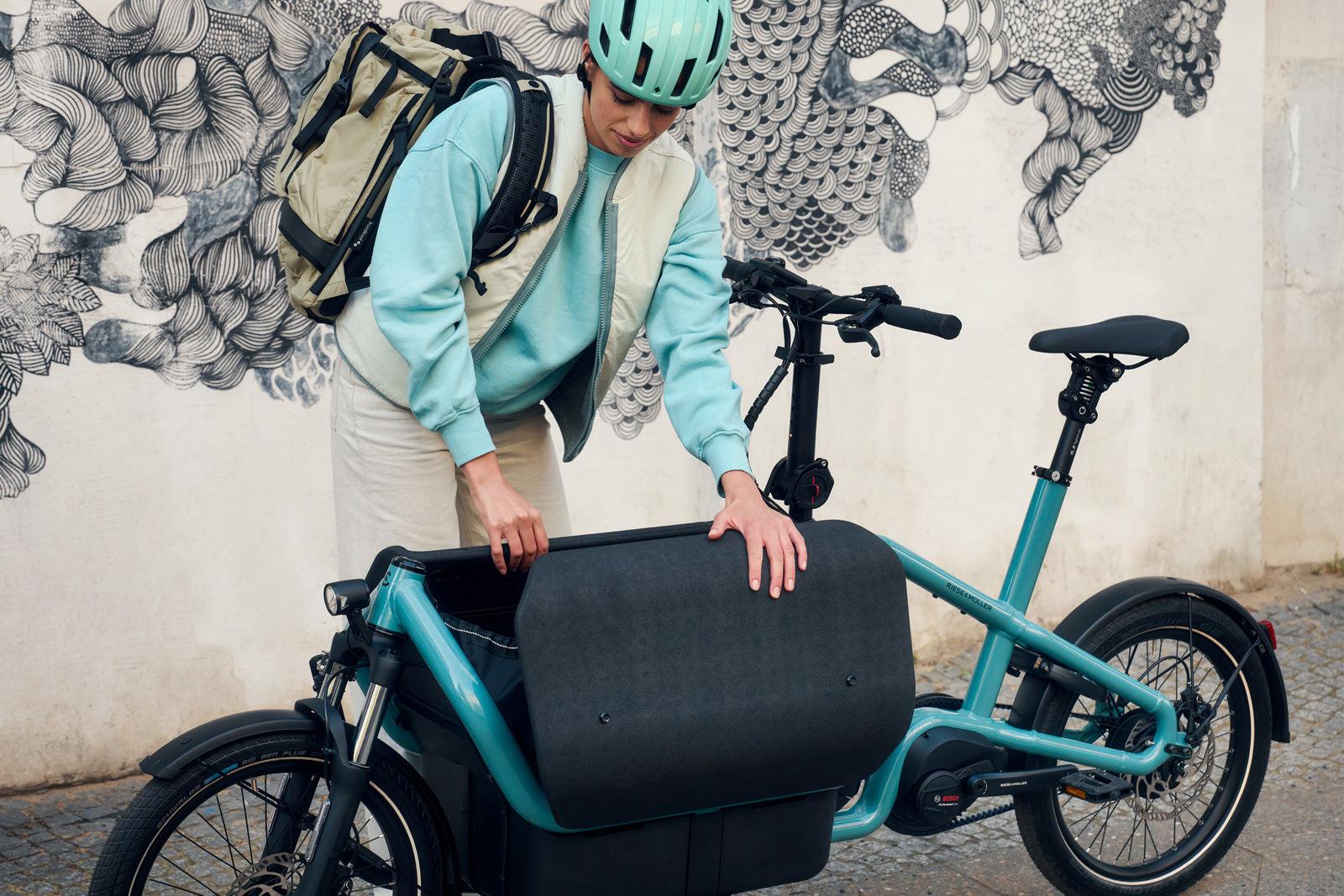 Helmeted cyclist adjusting the front cargo bag on a turquoise Riese & Müller electric cargo bike beside a mural-covered wall.