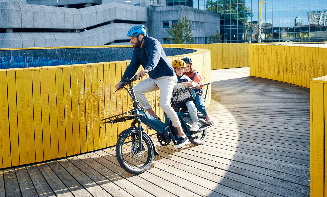 Man riding an electric cargo bike with two helmeted children on the rear seat along a wooden walkway with bright yellow railings.