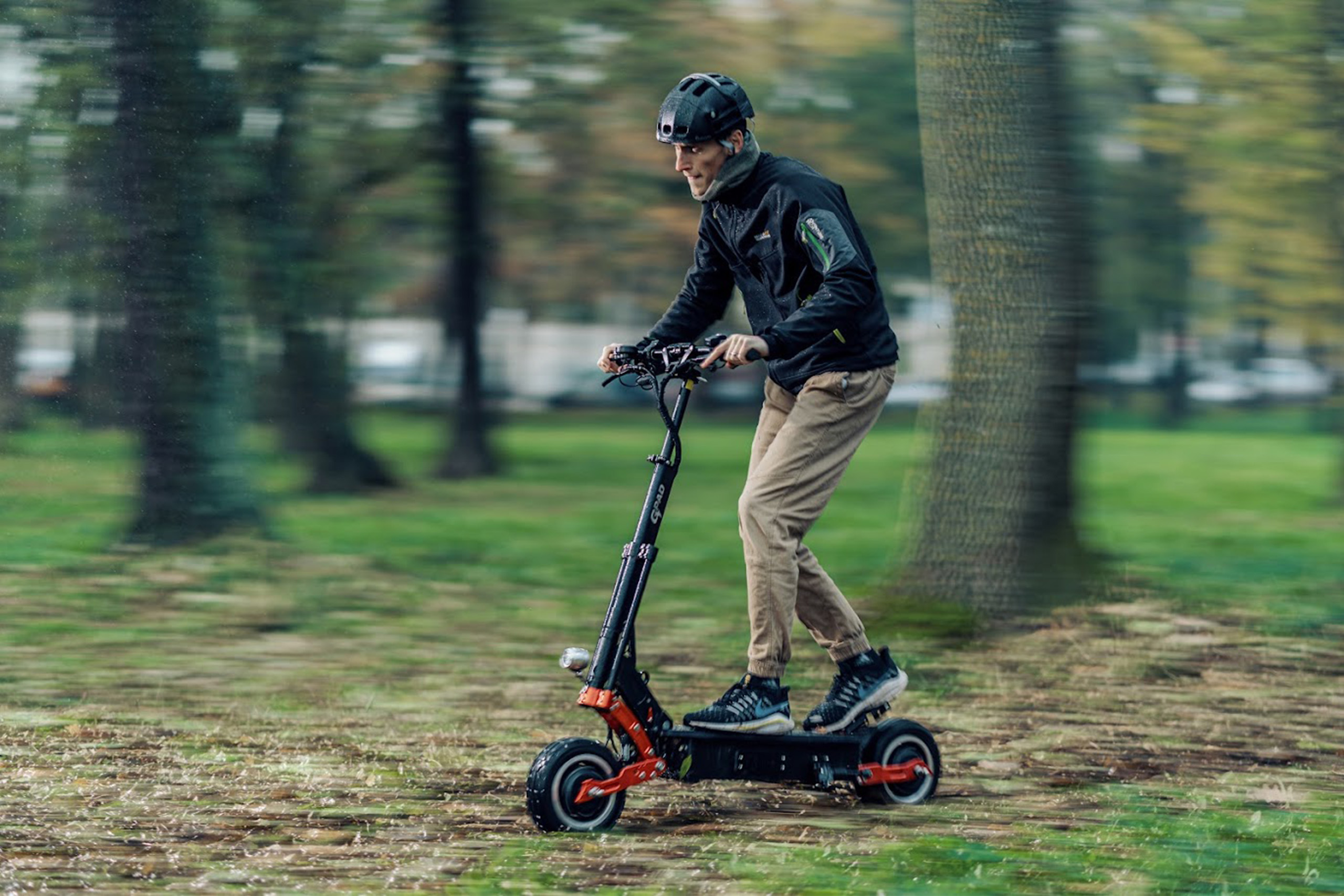 Electric scooter range forest blog image Helmeted man riding a powerful electric scooter through a green park with trees in the background.