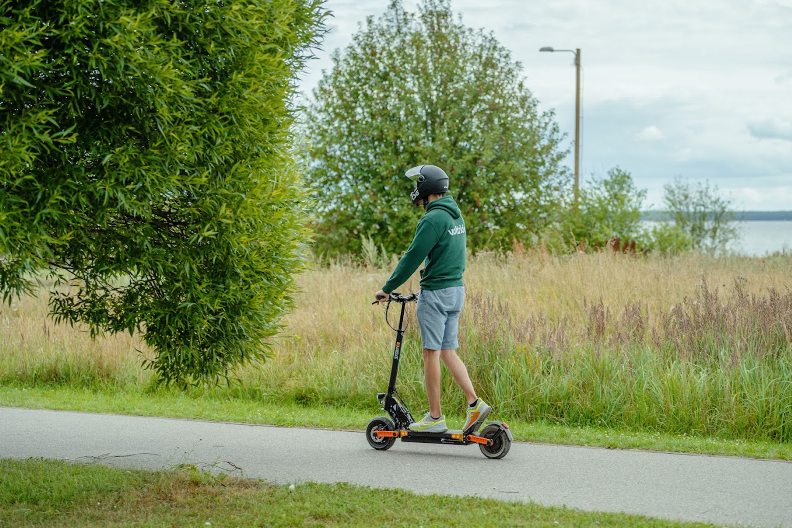 Electric scooter range blog image Helmeted man riding an electric scooter along a paved path beside a grassy field and lake.