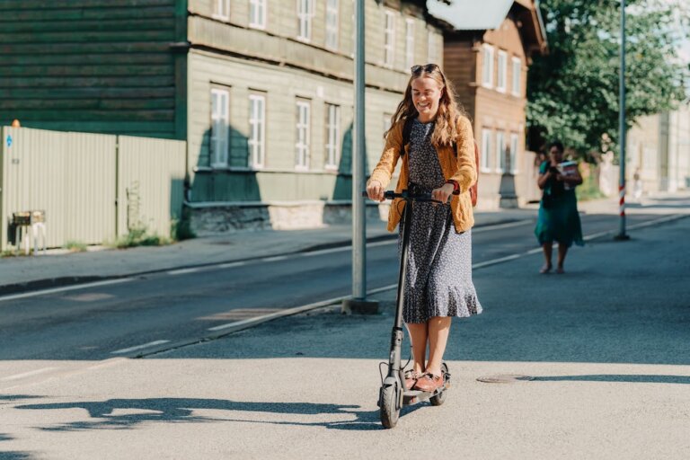 Woman riding an electric scooter on a sunny urban street lined with wooden buildings.