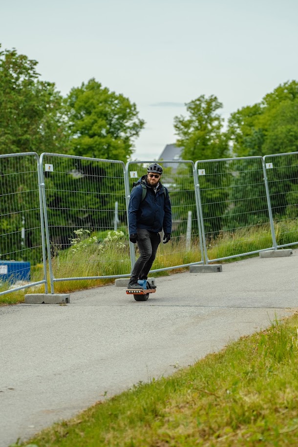Man wearing a helmet riding a Onewheel on a paved path beside construction fencing and greenery.